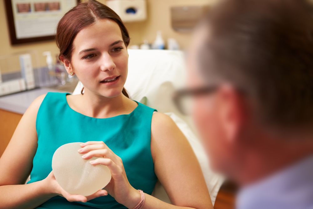 A woman wearing a turqoise shell top holding a breast implant with an inquisitive expression.